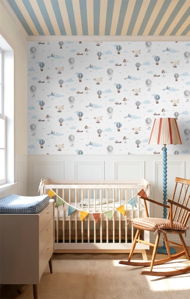 Helles Kinderzimmer mit Tapete mit Heißluftballons, Flugzeugen und Wolken über weißen Wandpaneelen, mit Babybett aus Holz und Schaukelstuhl.