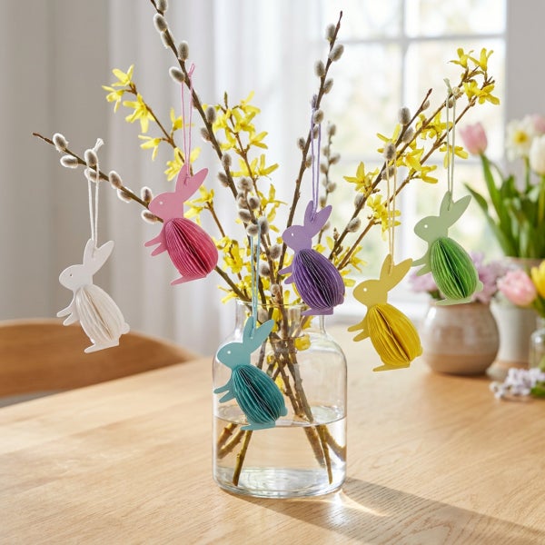 Osterhasen zum Aufhängen aus Papier als Dekoration an Weidenkätzchenzweigen und Forsythien in einer Glasvase auf einem Holztisch.