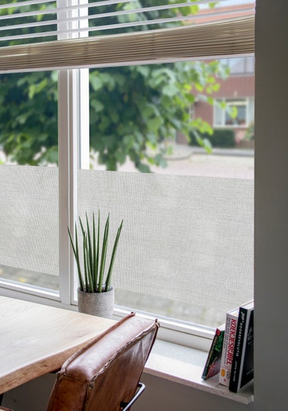 Halbtransparente Fensterfolie in Leinenoptik auf einer Fensterscheibe in einem hellen Raum mit Zimmerpflanze und Holztisch.