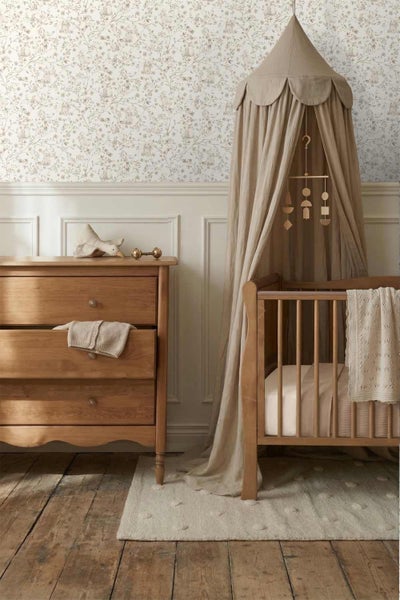 Vliestapete mit floralem Waldmotiv in einem Kinderzimmer mit Holzkommode, Gitterbett und Stoffhimmel über einem Holzdielenboden.