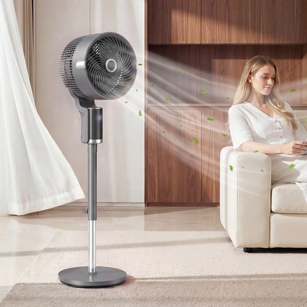 Moderner grauer Standventilator mit Digitalanzeige in hellem Wohnzimmer, Luftstrom ist auf eine sitzende Frau auf einem Sofa gerichtet.