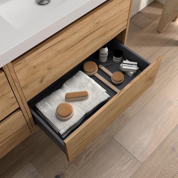 Offene Schublade eines Waschbeckenunterschranks in Eichenoptik mit Handtuch, Körperbürsten und Pflegeprodukten, organisiert und modern.