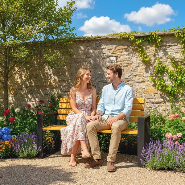 Ein Paar sitzt auf einer Gartenbank aus Holz mit Metallgestell vor einer Natursteinmauer in einem hellen Garten mit Blumen.