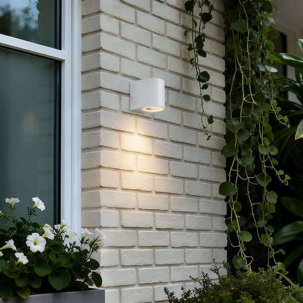 Moderne weiße Außenwandleuchte an einer hellen Ziegelwand neben einem Fenster und grünen Kletterpflanzen, Lichtstrahl nach unten gerichtet.