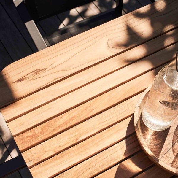 Detailansicht eines Gartentischs aus Holzlamellen mit einer Wasserflasche auf einem Holztablett im hellen Sonnenlicht.