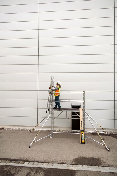 Eine Person mit Schutzhelm und Sicherheitsweste auf einem Gerüst mit einer Leiter an einer Gebäudewand.