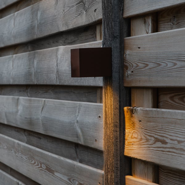 Rechteckige Außenwandleuchte aus Metall in Rostoptik, die warmes Licht nach unten auf einen Holzzaun wirft.