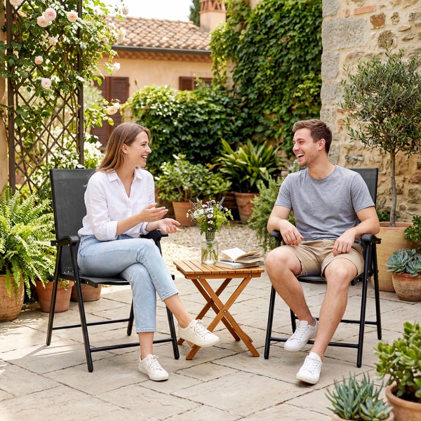 Ein Mann und eine Frau sitzen auf schwarzen Gartenstühlen an einem kleinen Klapptisch aus Holz auf einer Terrasse mit vielen Pflanzen.