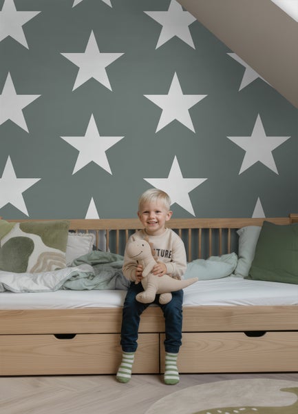 Kinderzimmer mit Tapete im Sterndesign, einem Holzbett mit Schubladen und Kissen. Ein Kind sitzt auf dem Bett und hält ein Stofftier.