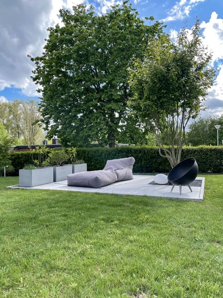 Moderne Sitzecke im Garten mit grauer Liege, schwarzem Schalensessel und rechteckigen Pflanzkübeln auf hellen Bodenplatten unter einem Baum.