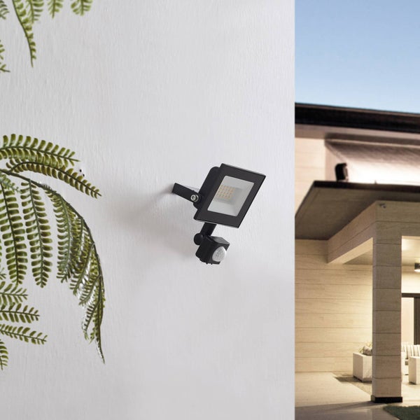 Schwarzer Außenstrahler mit Leuchtdioden und Bewegungsmelder an weißer Wand neben grünen Blättern mit Blick auf eine helle moderne Terrasse.