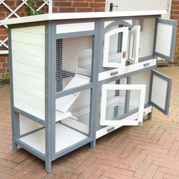 Zweistöckiger Kleintierstall aus Holz mit Rampe, mehreren Gittertüren und Holztüren auf einer gepflasterten Fläche.