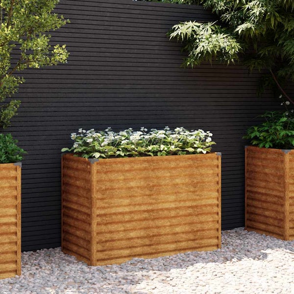 Rechteckiges Hochbeet in Rostoptik mit Wellenstruktur, bepflanzt mit Blumen vor einer dunklen Gartenwand auf hellem Kies.