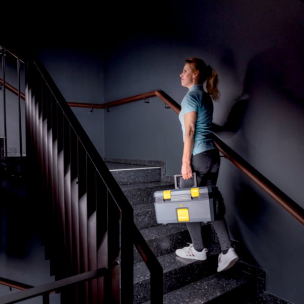Eine Frau trägt einen grauen Werkzeugkoffer mit gelben Verschlüssen eine Treppe in einem modernen Treppenhaus hinauf.