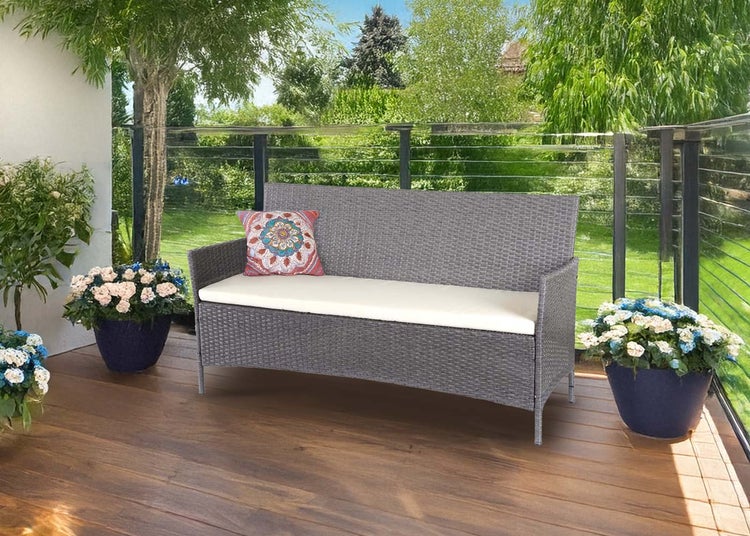 Graue Gartenbank aus Polyrattan auf einer Holzterrasse mit hellem Sitzkissen und buntem Zierkissen vor einem grünen Garten.