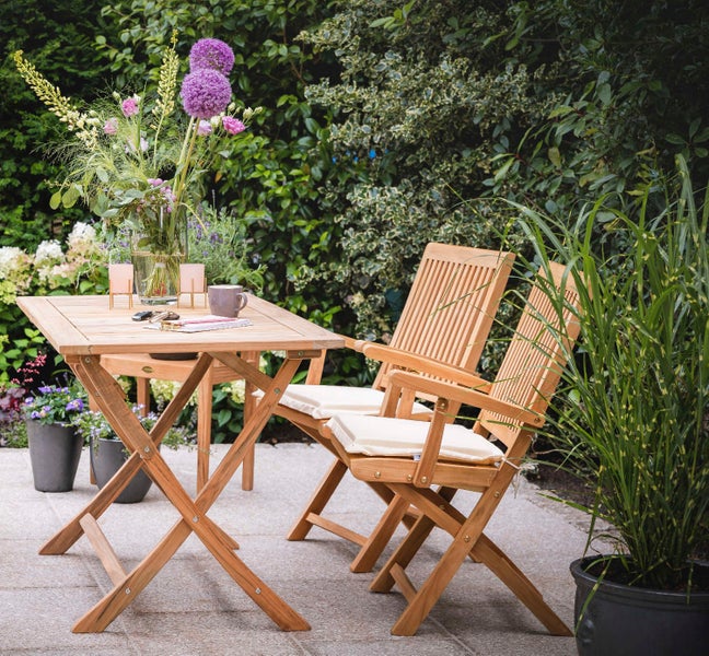 Gartenszene mit Holztisch und zwei Holzstühlen mit Sitzkissen auf einer Terrasse.