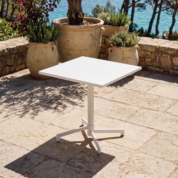 Quadratischer, weißer Bistrotisch auf einer hellen Steinterrasse mit großen Terrakotta-Pflanzkübeln und Küstenpanorama.