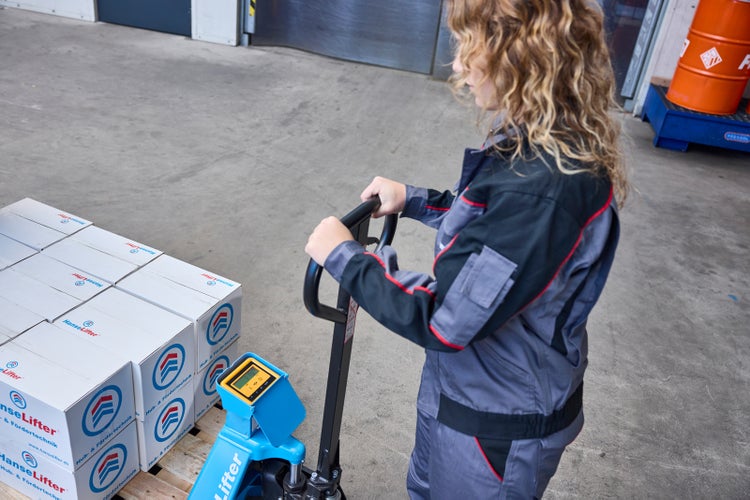 Eine Person bedient einen Hubwagen mit Waage und digitaler Anzeige beim Transport einer Palette mit Kartons.