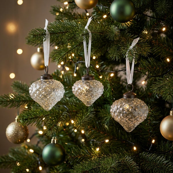 Drei herzförmige Weihnachtskugeln aus Glas mit strukturierter Oberfläche, hängend an einem Weihnachtsbaum mit Lichterkette und Weihnachtsschmuck.