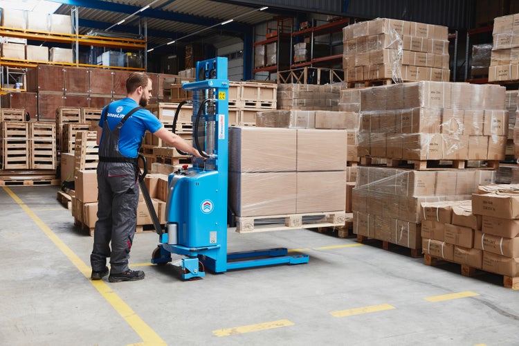 Ein blauer Elektro-Deichselstapler hebt eine Palette mit Kartons in einem Lager an, bedient von einer Person in Arbeitskleidung.