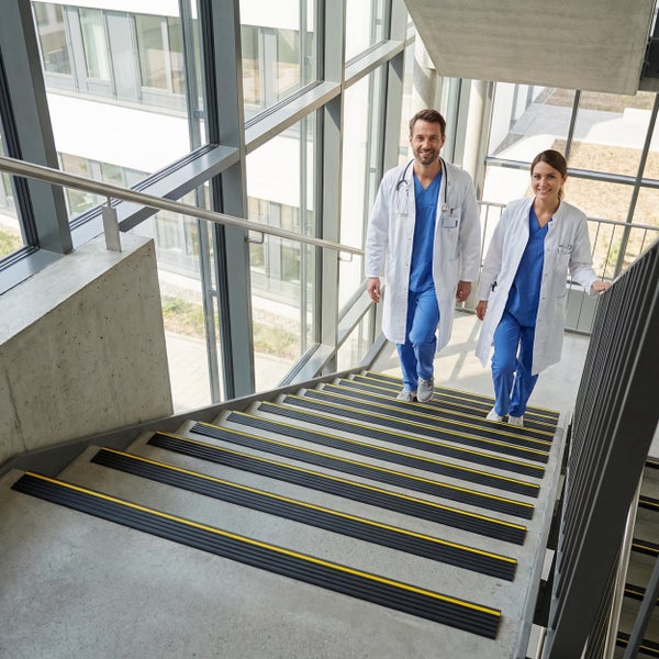 Zwei Mediziner in Kitteln gehen eine Betontreppe mit rutschhemmenden Kantenprofilen in einem modernen, lichtdurchfluteten Treppenhaus entlang.