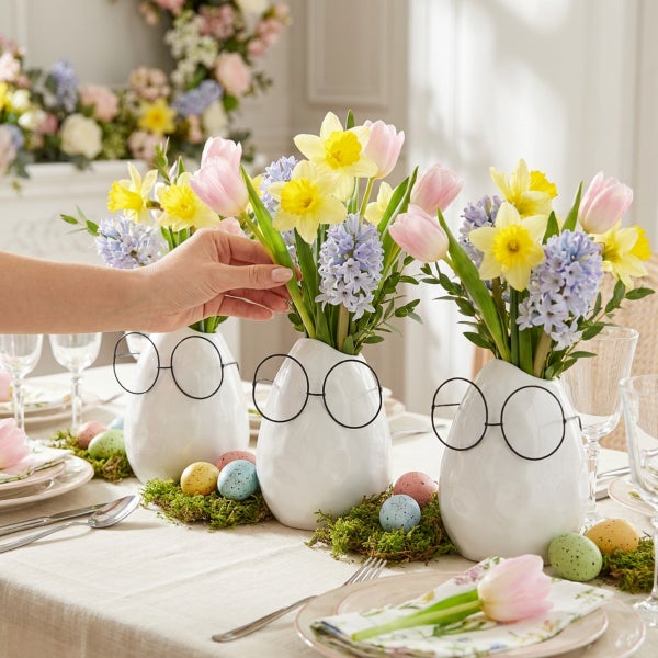 Drei weisse eiförmige Vasen mit schwarzem Brillengestell und Frühlingsblumen dekoriert auf einem gedeckten Ostertisch mit buntem Ei-Schmuck.