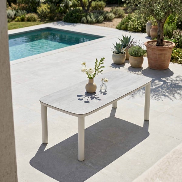 Moderner rechteckiger Gartentisch mit abgerundeten Ecken auf einer hellen Terrasse neben einem Pool, dekoriert mit Blumenvasen.