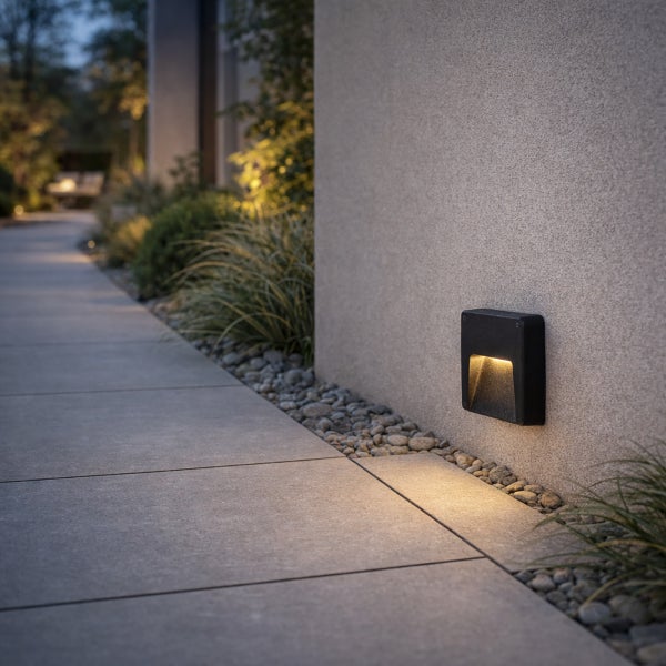Rechteckige schwarze LED Außenwandleuchte an einer verputzten Wand, die einen gepflasterten Gartenweg bei Dunkelheit beleuchtet.