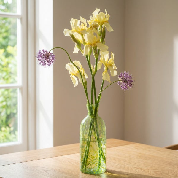 Eine Glasvase mit gelben Schwertlilien und violetten Zierlauchblüten auf einem Holztisch vor einem Fenster