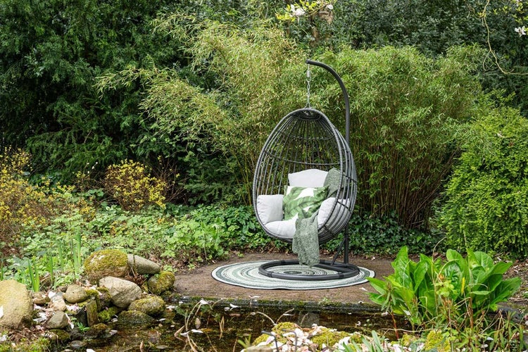 Hängesessel in Eiform mit Gestell, graues Geflecht, Polster und Decke, in einem grünen Garten mit Teich und Pflanzen.