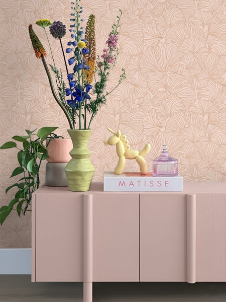 Wohnraum mit gemusterter Blumentapete, einem rosa Sideboard und Dekoelementen wie einer Blumenvase und einer Einhornfigur.