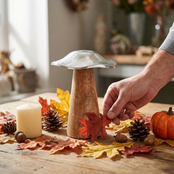 Herbstliche Tischdekoration mit einer Pilzfigur aus Holz und Metall, umgeben von Blättern, Tannenzapfen, einer Kerze und einem kleinen Zierkürbis.