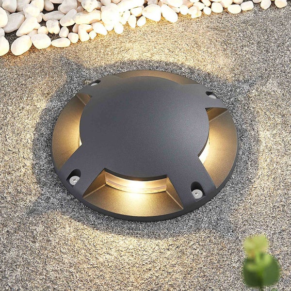 Runde Bodeneinbauleuchte mit vier seitlichen Lichtauslässen, in Kies eingebettet, strahlt warmweißes Licht aus.