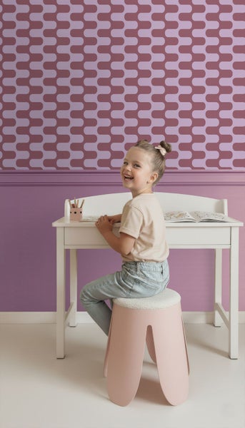 Ein Mädchen sitzt an einem weißen Schreibtisch auf einem Hocker in einem Kinderzimmer mit gemusterter Tapete.