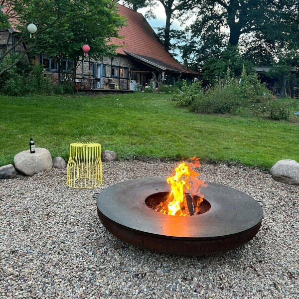 Große kreisförmige Feuerschale aus Stahl mit breitem Grillring und brennendem Holzfeuer auf Kiesfläche in einem weitläufigen Garten mit Rasen.