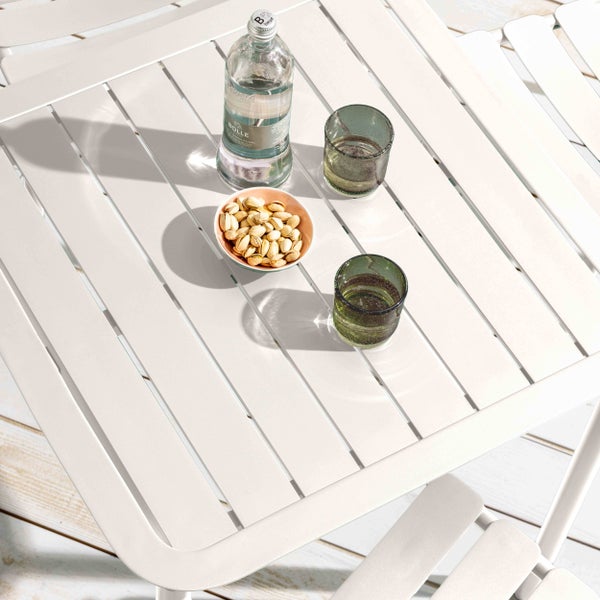 Weißer Gartentisch mit Lattenoberfläche aus Metall, darauf eine Flasche Wasser, zwei Gläser und eine Schale Pistazien.