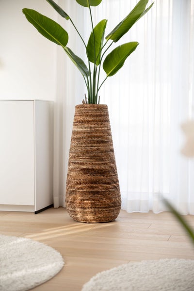 Hoher geflochtener Pflanzkübel aus Naturmaterial mit einer großblättrigen Zimmerpflanze in einem hellen, modernen Wohnraum.