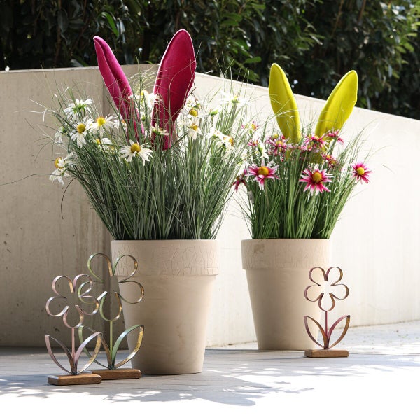 Zwei Pflanzentöpfe mit Osterhasenohren, Blumen und Gras sowie dekorative Metallblumen auf einem Holzsockel als Frühlingsdekoration.
