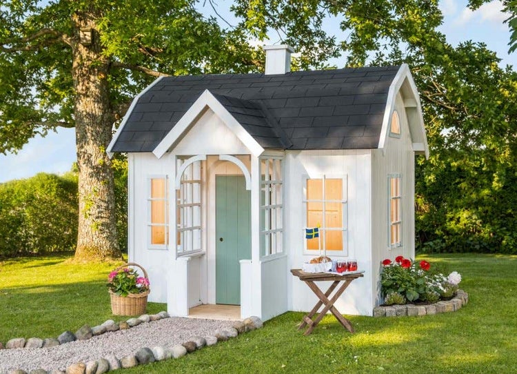 Weißes Spielhaus aus Holz mit dunklem Schindeldach, kleiner Veranda und grüner Tür in einem hellen Garten.