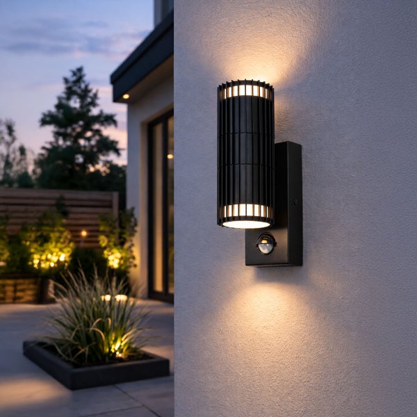 Moderne schwarze zylindrische Außenwandleuchte mit Bewegungsmelder, Up-and-Down-Lichteffekt an einer Hauswand im Garten bei Dämmerung.