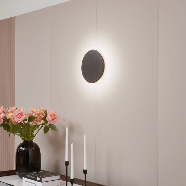 Runde Wandleuchte in Schieferoptik mit indirekter Beleuchtung in einem hellen Raum mit Blumen und Kerzen.