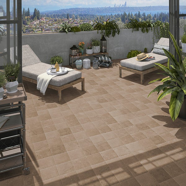 Terrasse mit sandfarbenen Bodenfliesen in Steinoptik, eingerichtet mit zwei Liegen, Pflanzen und einem Beistelltisch vor städtischem Ausblick.