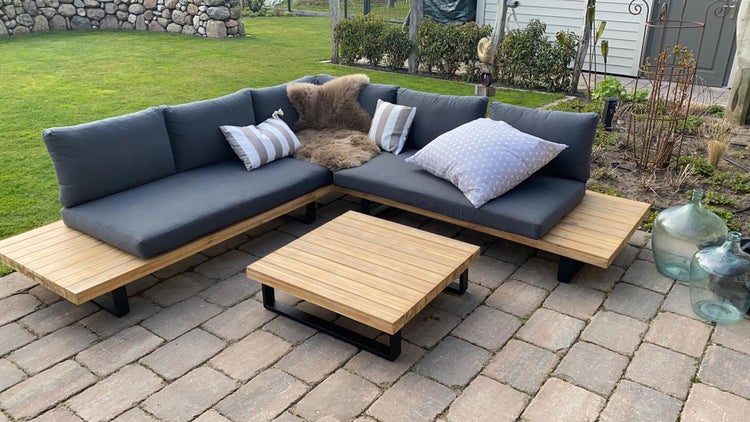 Modernes Lounge-Set aus Holz mit grauem Ecksofa und quadratischem Couchtisch auf einer gepflasterten Terrasse im Garten.