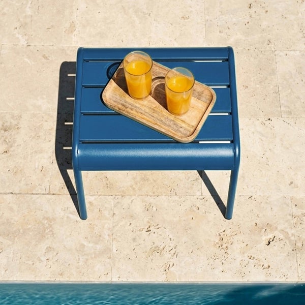Blauer Garten-Beistelltisch aus Metall mit Holztablett und zwei Gläsern Orangensaft auf einer Steinterrasse neben einem Pool.