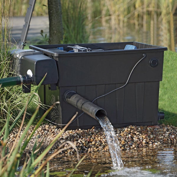Schwarzes Teichfiltersystem am Rand eines Gartenteichs, aus dem Wasser über ein Rohr zurück in den Teich fließt.