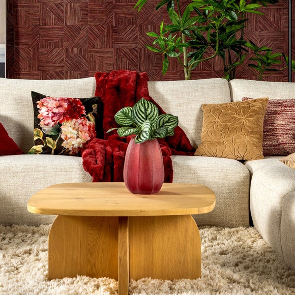 Modernes Wohnzimmer mit hellem Sofa, Holz-Couchtisch, gemusterten Kissen, grüner Pflanze und roter geometrischer Tapete.