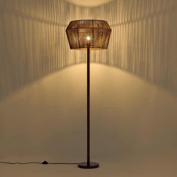 Stehleuchte mit einem Schirm aus feinem Geflecht, die ein warmes Licht und dekorative Streifen-Schatten an die Wand wirft.