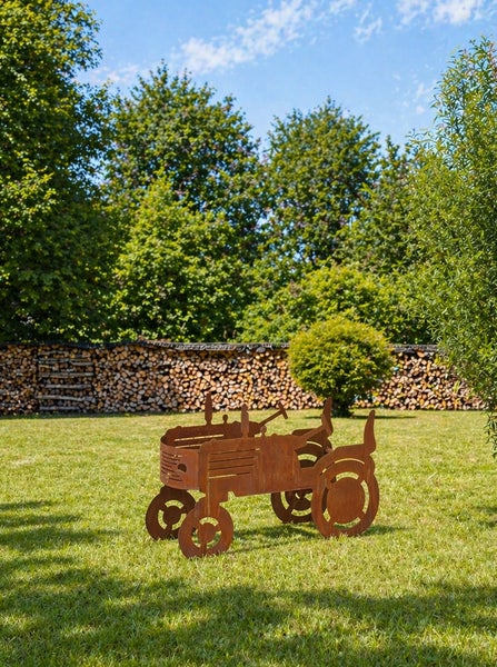 Rostige Garten-Dekoration in Form eines Traktors auf einer Wiese vor einem Holzlager und Bäumen.