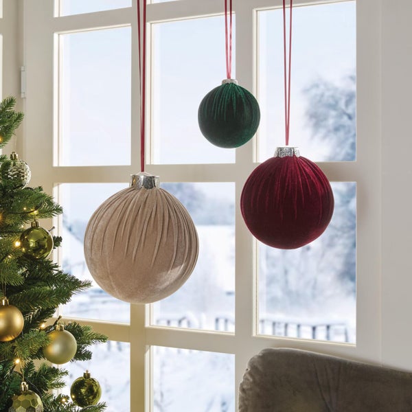 Drei hängende Weihnachtskugeln aus Samt vor einem Fenster, daneben ein beleuchteter Weihnachtsbaum.