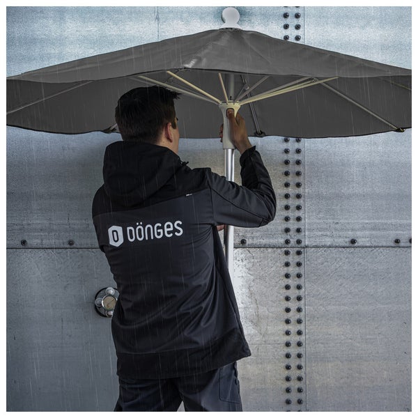 Eine Person in einer Jacke mit Dönges Logo steht unter einem großen grauen Regenschirm im Regen vor einer Metallwand.